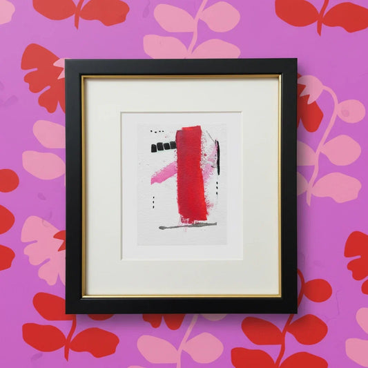 Framed mini watercolor painting with red, pink, and black abstract shapes on floral pink wall.