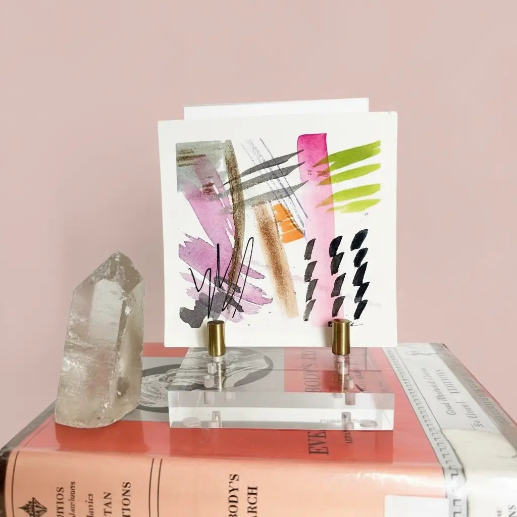 Two perfume bottles on a clear stand with abstract art card on a pink surface