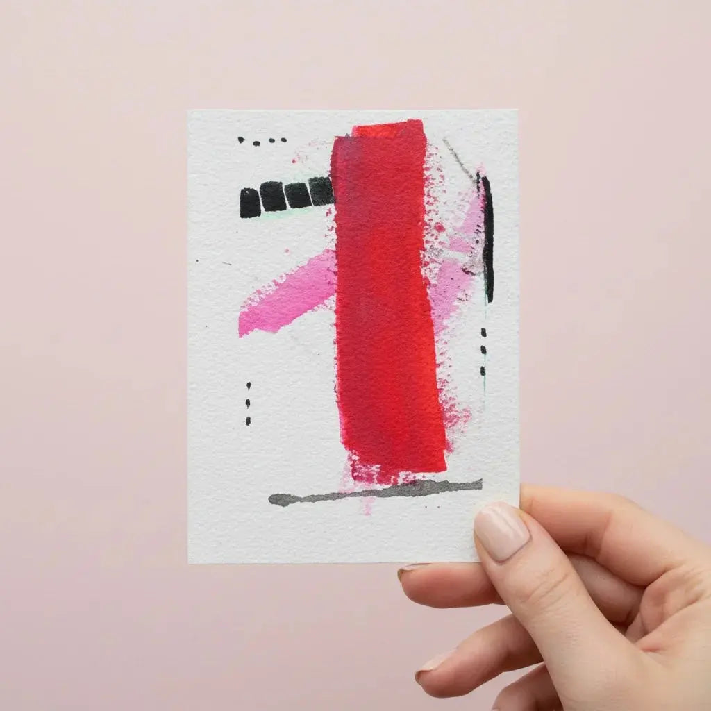 Hand holding a mini abstract watercolor painting with red, pink, and black brushstrokes